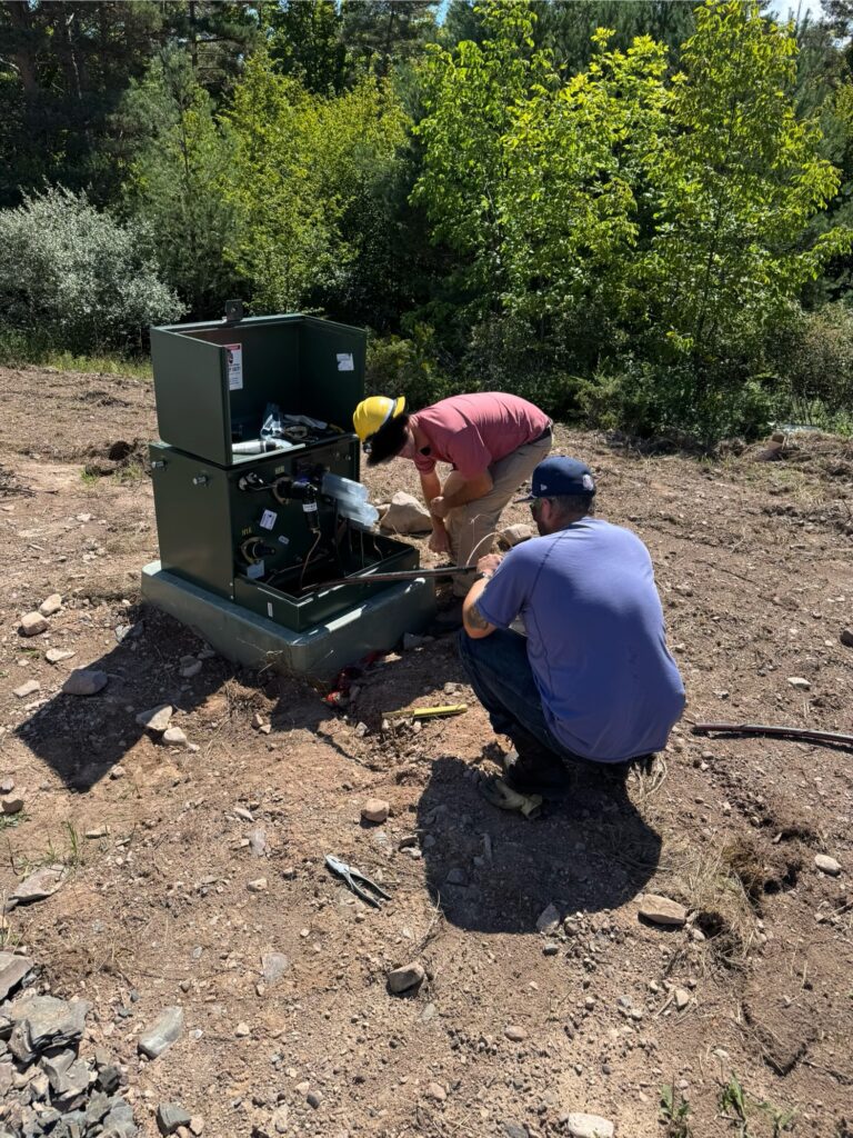 Two utility workers kneel on the ground beside an open green electrical box while installing or repairing equipment on a rural property, surrounded by dirt, rocks, and trees under bright sunlight.