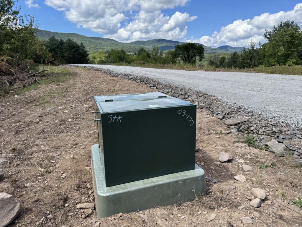 A newly installed green electrical utility box sits on a gravel and dirt roadside with mountains and trees in the background under a bright blue sky with clouds.